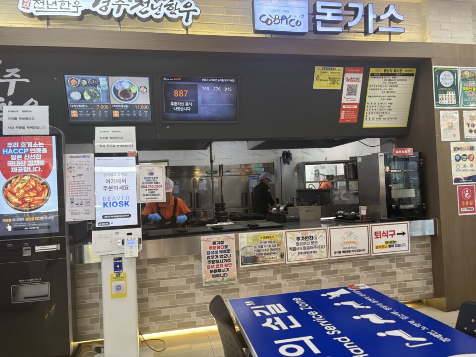 フードコートの韓牛クッパ・トンカツ店。番号札の呼び出しモニターとキオスク