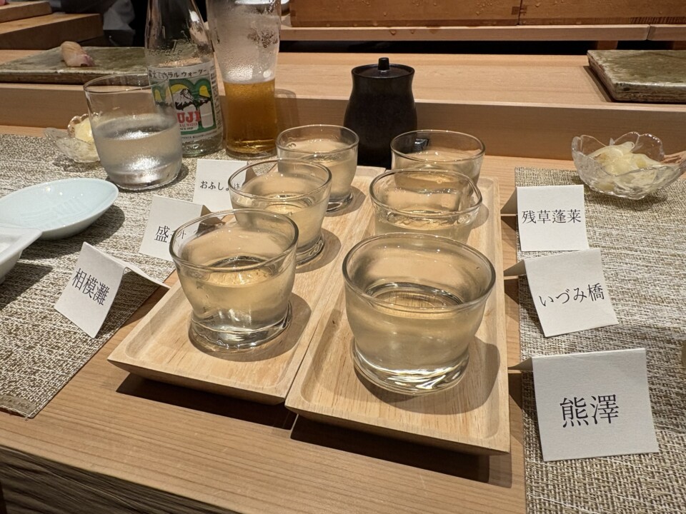 Six small glasses of sake served on wooden trays with cards naming each brewery
